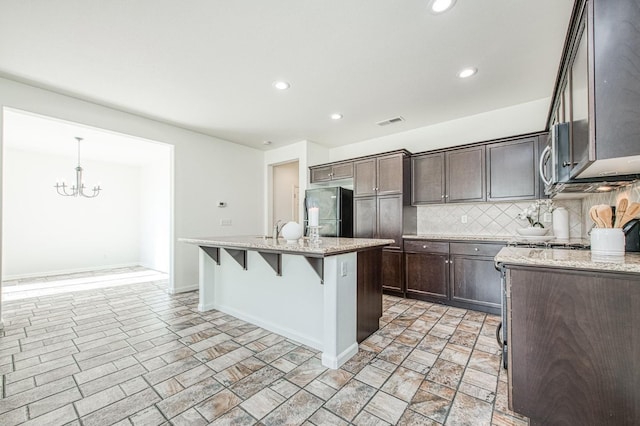 kitchen with visible vents, decorative backsplash, freestanding refrigerator, an island with sink, and light stone countertops