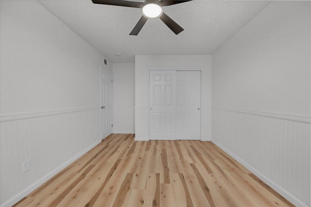 unfurnished bedroom with a wainscoted wall, a closet, light wood-style floors, a ceiling fan, and a textured ceiling