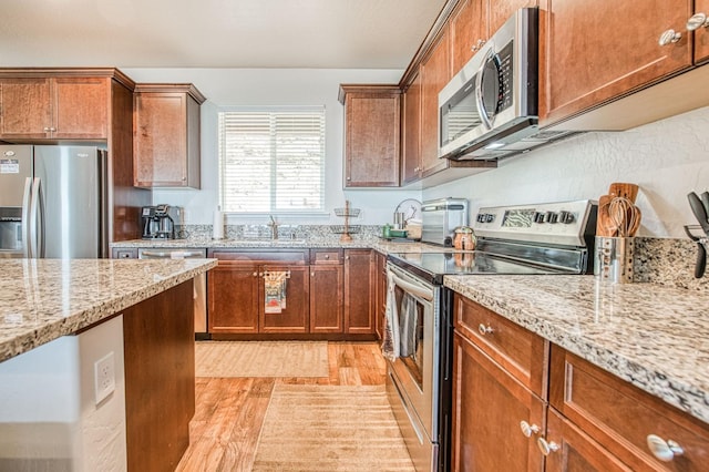 kitchen featuring appliances with stainless steel finishes, brown cabinets, light stone countertops, light wood-type flooring, and a sink