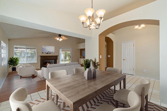dining room featuring light tile patterned flooring, vaulted ceiling, a lit fireplace, arched walkways, and ceiling fan with notable chandelier