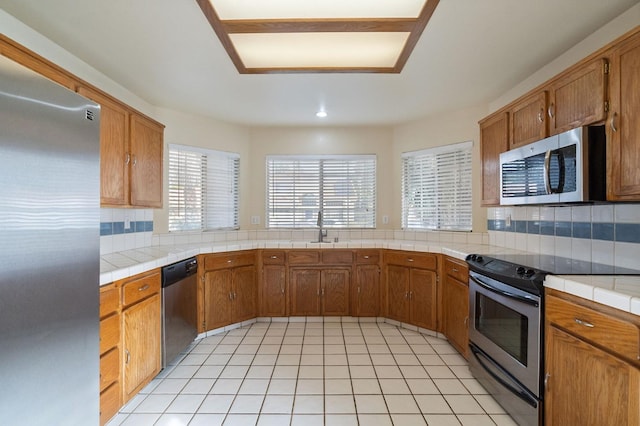 kitchen featuring tile countertops, brown cabinetry, appliances with stainless steel finishes, and decorative backsplash