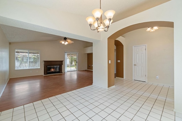 unfurnished living room featuring arched walkways, light tile patterned flooring, ceiling fan with notable chandelier, vaulted ceiling, and a glass covered fireplace