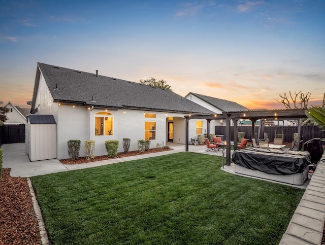 back of house at dusk featuring fence private yard, a yard, roof with shingles, stucco siding, and a patio area