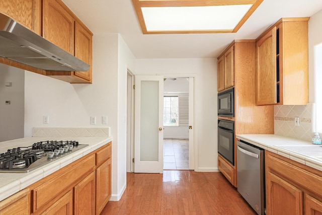 kitchen featuring tile counters, decorative backsplash, light wood-type flooring, black appliances, and exhaust hood