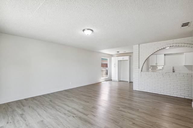 unfurnished living room featuring baseboards, visible vents, brick wall, wood finished floors, and a textured ceiling