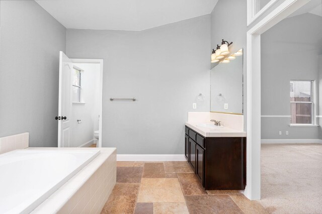 bathroom featuring a relaxing tiled tub, baseboards, toilet, stone finish floor, and vanity