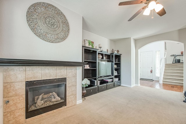 carpeted living room featuring baseboards, arched walkways, a ceiling fan, a tiled fireplace, and stairs