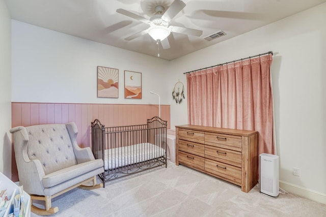 carpeted bedroom with a nursery area, a wainscoted wall, visible vents, and ceiling fan