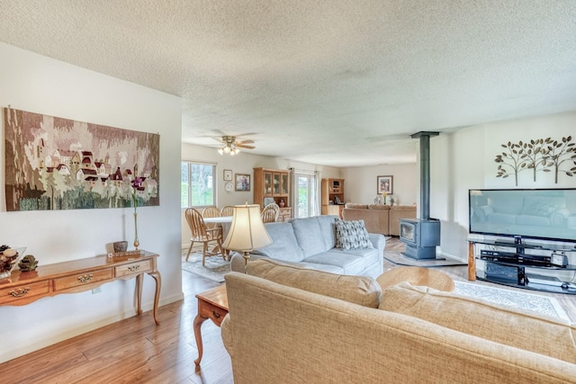 living room featuring a textured ceiling, a ceiling fan, baseboards, light wood finished floors, and a wood stove