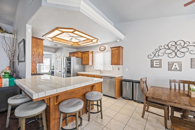 kitchen with light tile patterned floors, tile counters, brown cabinetry, appliances with stainless steel finishes, and a peninsula