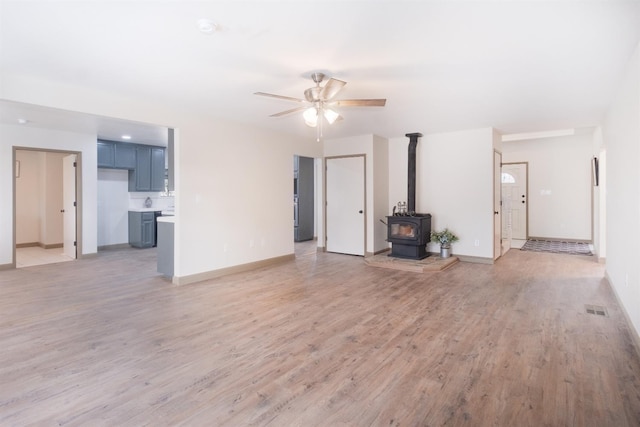 unfurnished living room featuring light wood finished floors, baseboards, visible vents, a ceiling fan, and a wood stove