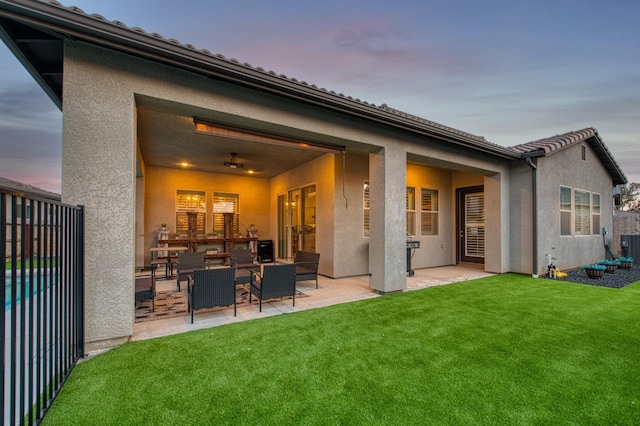 back of property at dusk with a ceiling fan, a patio, fence, a yard, and stucco siding