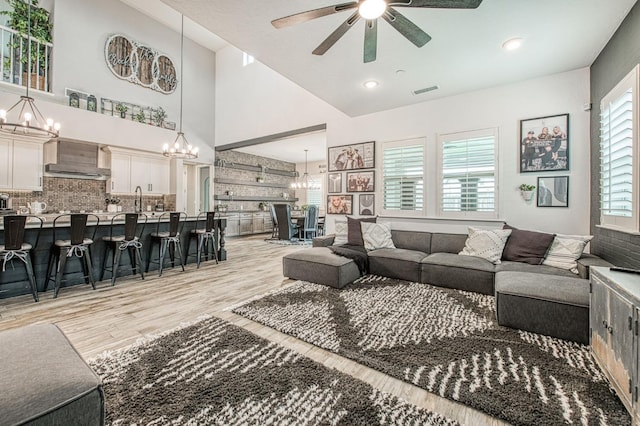 living room featuring a towering ceiling, light wood-style flooring, visible vents, and ceiling fan with notable chandelier