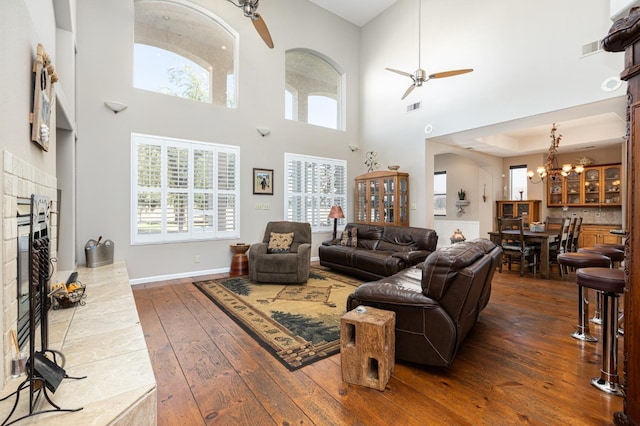 living area with a fireplace with raised hearth, ceiling fan with notable chandelier, visible vents, baseboards, and dark wood-style floors