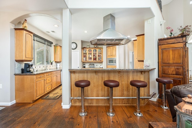 kitchen featuring island exhaust hood, dark wood finished floors, stainless steel microwave, a kitchen bar, and glass insert cabinets