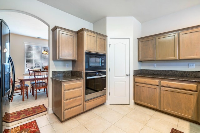 kitchen with arched walkways, light tile patterned floors, light carpet, dark stone counters, and black appliances