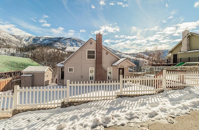 snow covered house featuring an outbuilding, a chimney, a mountain view, a shed, and fence private yard