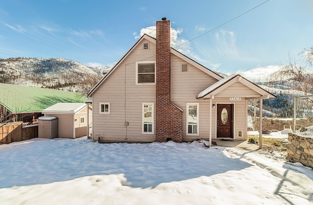 snow covered rear of property with a mountain view, a chimney, an outdoor structure, and a storage shed