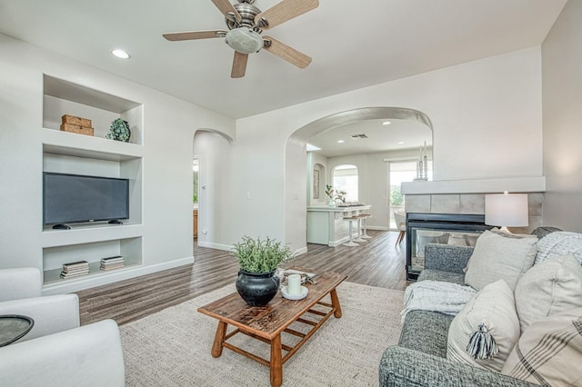 living area with baseboards, built in features, arched walkways, ceiling fan, and wood finished floors