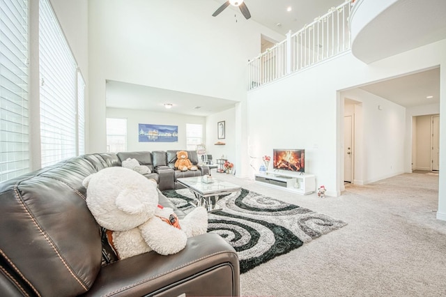 living room with baseboards, a ceiling fan, a towering ceiling, a lit fireplace, and carpet flooring