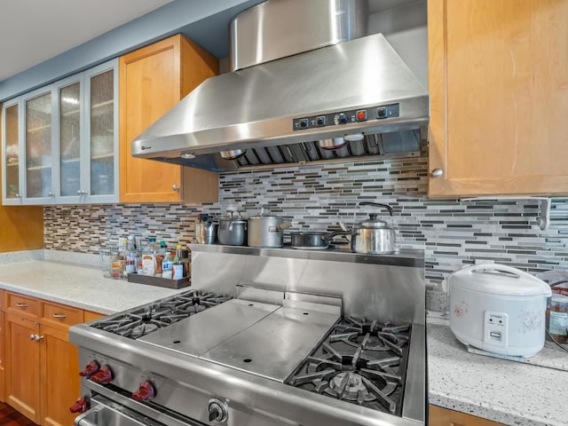 kitchen featuring wall chimney range hood, backsplash, light stone countertops, gas stove, and glass insert cabinets