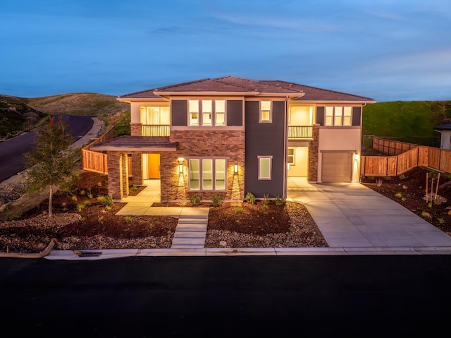 prairie-style home with an attached garage, a balcony, fence, stone siding, and concrete driveway