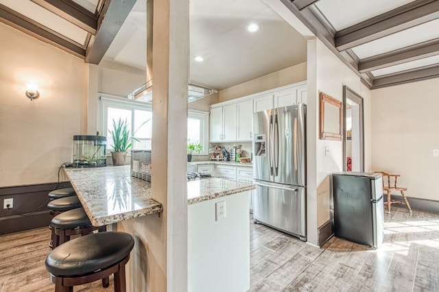 kitchen with light wood-style flooring, stainless steel appliances, white cabinets, light stone countertops, and beamed ceiling