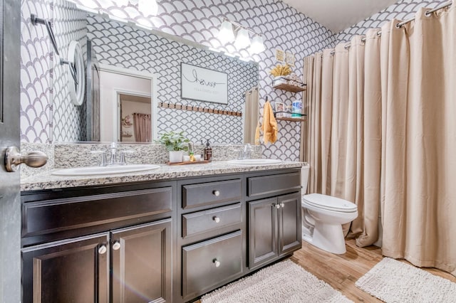 full bath with double vanity, a sink, toilet, and wallpapered walls