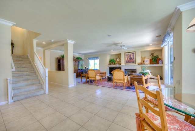 dining space featuring crown molding, light tile patterned floors, ceiling fan, a tile fireplace, and stairs