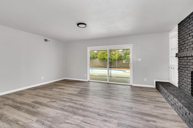 unfurnished living room with a textured ceiling, wood finished floors, visible vents, baseboards, and a brick fireplace