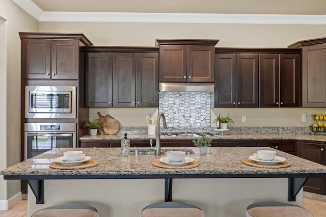 kitchen featuring dark brown cabinetry, a breakfast bar area, stainless steel appliances, crown molding, and under cabinet range hood