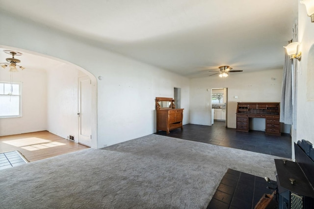 unfurnished living room with arched walkways, ceiling fan, dark carpet, and visible vents