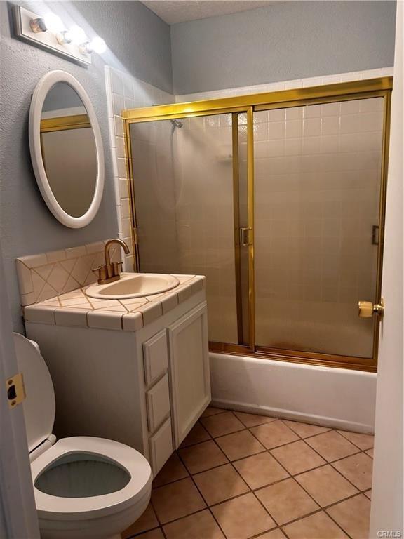 full bath with a textured wall, toilet, combined bath / shower with glass door, vanity, and tile patterned flooring