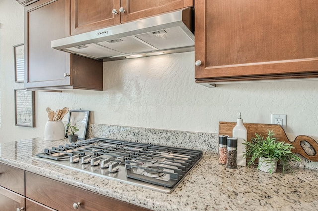 kitchen with a textured wall, under cabinet range hood, stainless steel gas cooktop, light stone countertops, and brown cabinetry