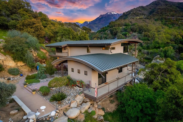 back of house at dusk with metal roof, a mountain view, a balcony, stucco siding, and a standing seam roof