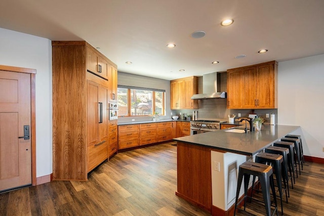 kitchen with dark wood-style floors, a breakfast bar, appliances with stainless steel finishes, wall chimney range hood, and a peninsula