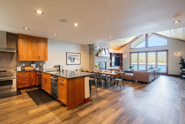 kitchen featuring a peninsula, wall chimney range hood, appliances with stainless steel finishes, and brown cabinets