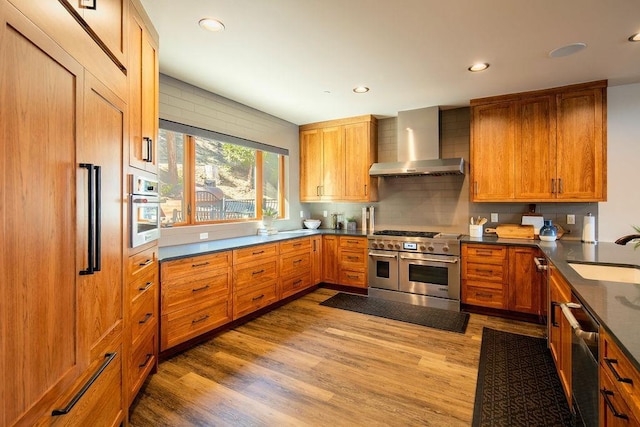 kitchen with dark countertops, wall chimney exhaust hood, light wood-style flooring, stainless steel appliances, and a sink