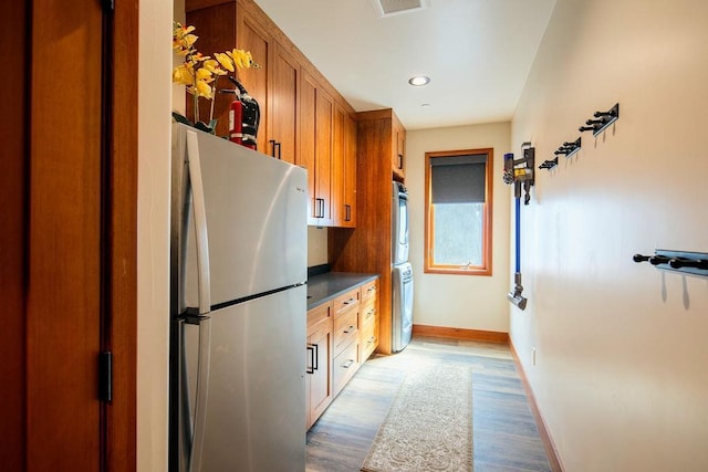 kitchen with dark countertops, brown cabinetry, freestanding refrigerator, stacked washing maching and dryer, and wood finished floors