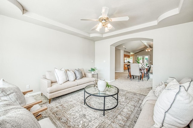 living room with carpet floors, arched walkways, a tray ceiling, a ceiling fan, and baseboards