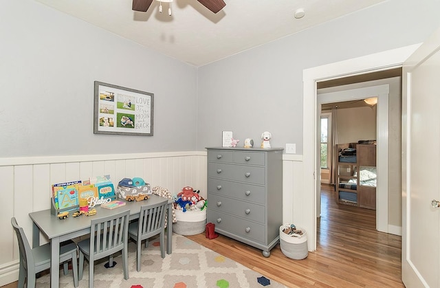 recreation room with light wood-type flooring, a wainscoted wall, and a ceiling fan