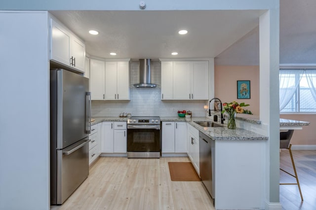 kitchen featuring appliances with stainless steel finishes, light wood-style floors, a sink, a peninsula, and wall chimney exhaust hood