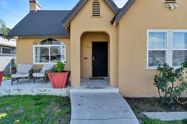 entrance to property featuring a shingled roof, a chimney, fence, and stucco siding