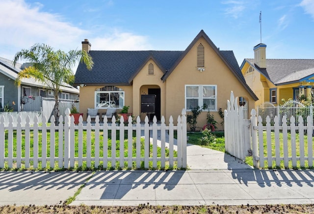 tudor house with roof with shingles, a fenced front yard, a chimney, and stucco siding