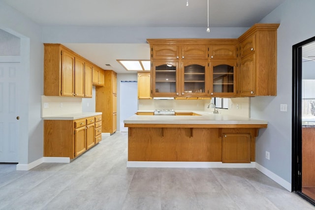 kitchen with a peninsula, a sink, light countertops, brown cabinetry, and glass insert cabinets