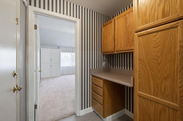 interior space featuring light carpet, wallpapered walls, baseboards, brown cabinets, and light countertops