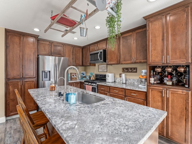 kitchen featuring stainless steel appliances, wood finished floors, a kitchen island with sink, a sink, and light stone countertops