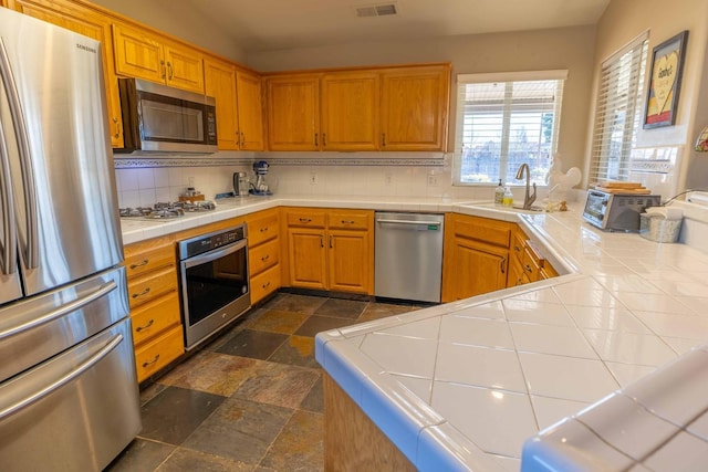 kitchen with stone tile floors, stainless steel appliances, tile counters, decorative backsplash, and a sink