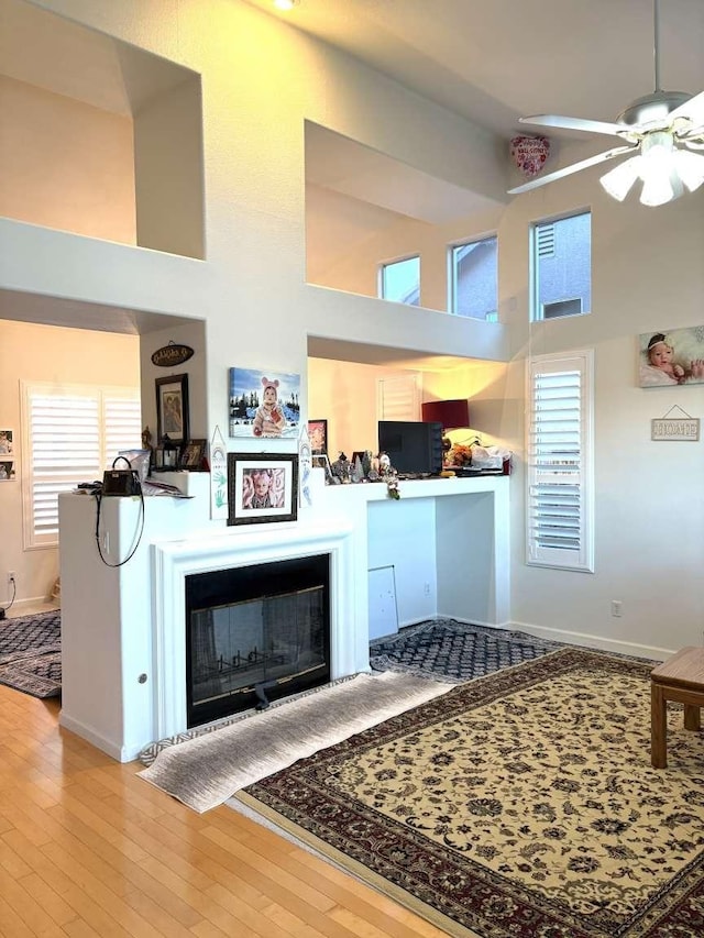 living area featuring baseboards, a glass covered fireplace, ceiling fan, hardwood / wood-style floors, and high vaulted ceiling