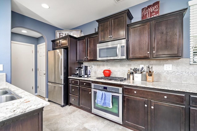 kitchen with light stone counters, arched walkways, stainless steel appliances, dark brown cabinets, and tasteful backsplash